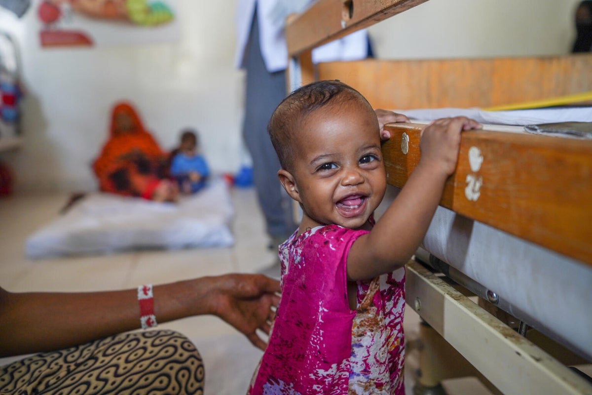A toddler girl holds onto a bed at a Stablisation Centre in Somalia while looking at the camera and smiling. 