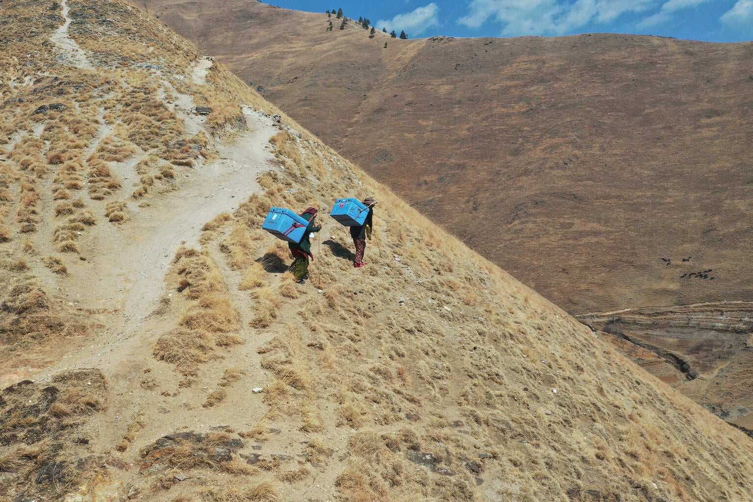 Two men are walking through a deserted mountain trail. They are carrying an cooler box on their bags with the UNICEF logo
