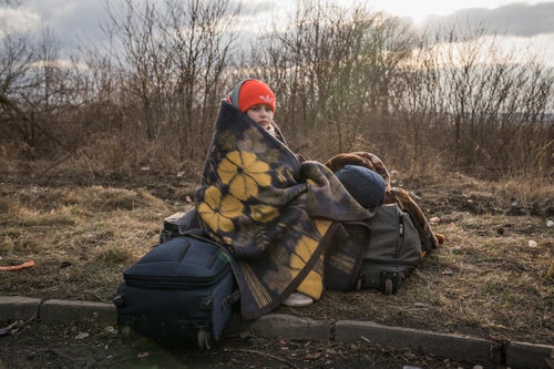 A young girl with blankets wrapped around her, sitting on luggage on the side of a road. 