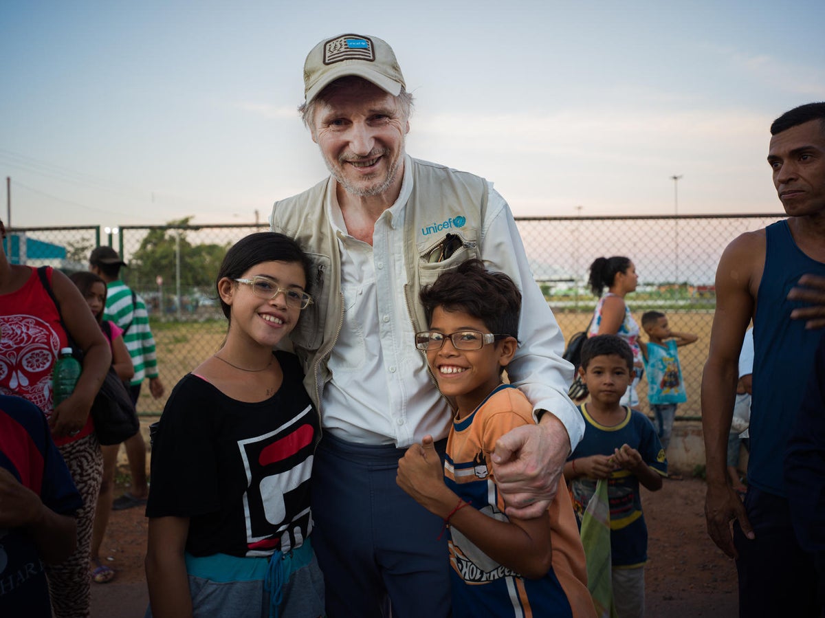 Liam Neeson poses with a young boy and a young girl. He is wearing a UNICEF vest.