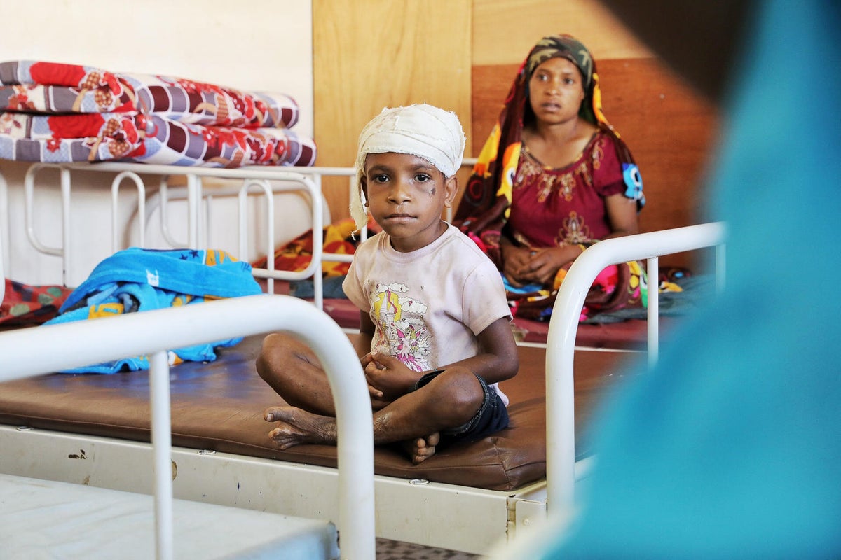 A boy is seating on a hospital bed. He has his head bandaged.