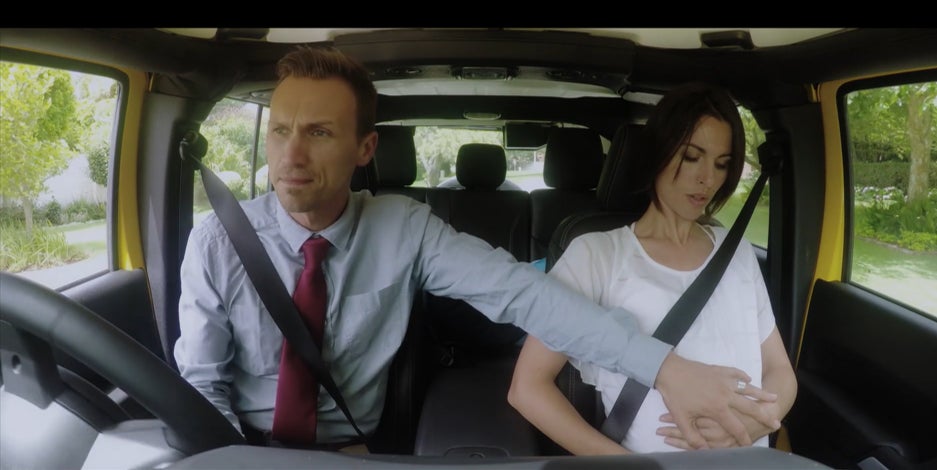 A man and a woman are seating in a car. The woman is pregnant and the man is driving, and has one hand on the woman's stomach.