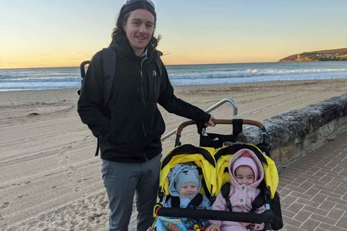 A man carrying a double pram with two children on a it. 