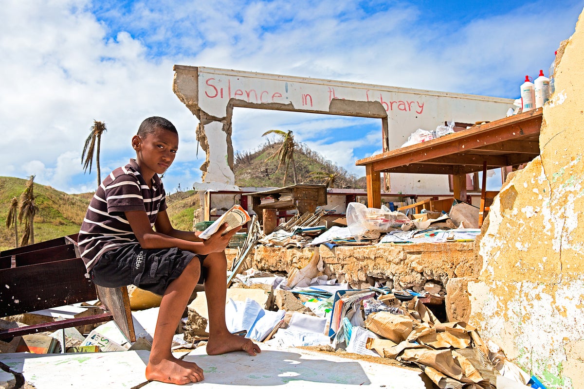 Another boy is seating in what looks like a room destroyed by a weather event.