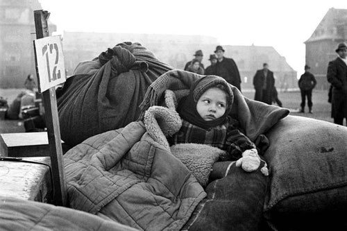 In 1946, inside Germany, a small displaced girl is wrapped in blankets and sits on a mound of other refugee belongings. 