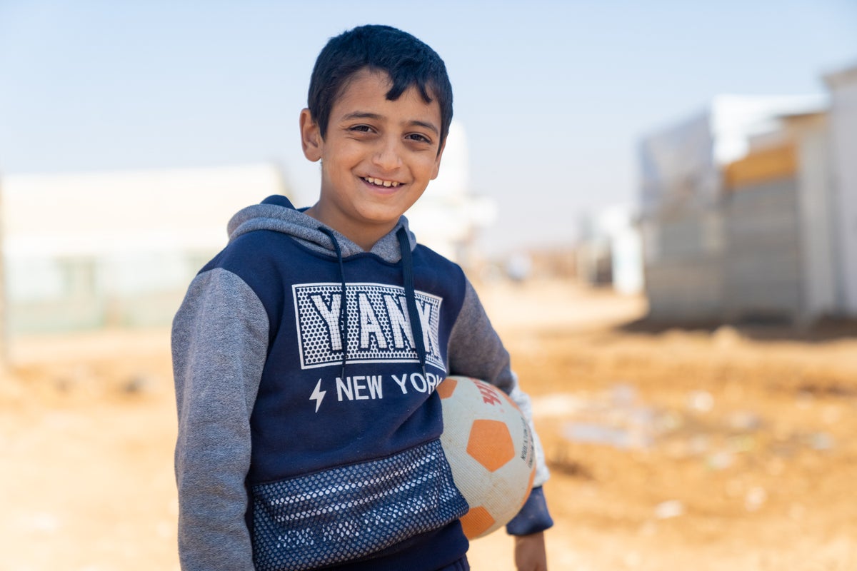 A Syrian boy in a refugee camp smiling at the camera while holding a soccer ball. 