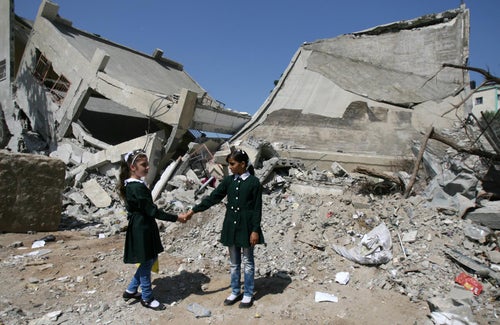 Two girls hold hands. In the background, a destroyed building