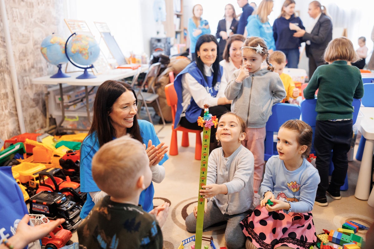 A smiling woman sitting at a children's desk surrounded by children. 