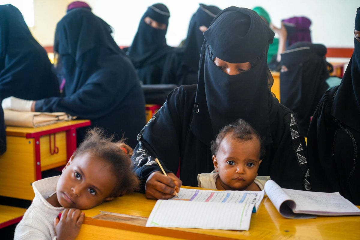 A woman writes in a notebook during a lesson as an infant child sits in her lap and an older child stands next to the desk. 