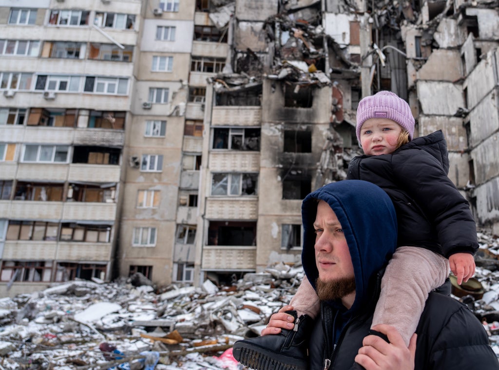 A father with his daughter on his shoulders standing outside a destroyed apartment building