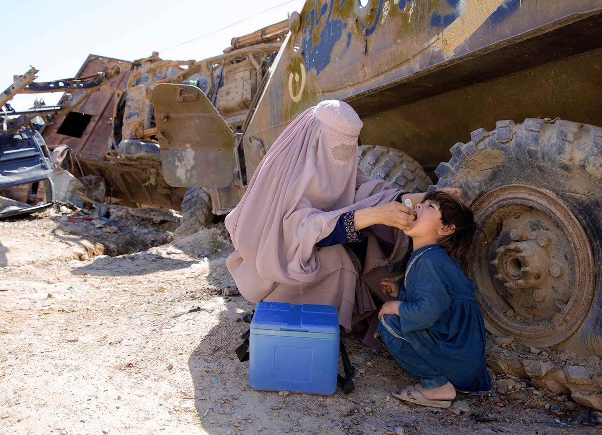 A woman wearing a burqa is giving a droplet vaccine to a boy. The boy is seating on the floor. Behind them there's a military vehicle.