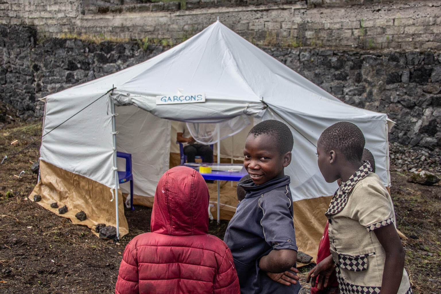 Young children looking at the camera in front of a makeshift tent. 