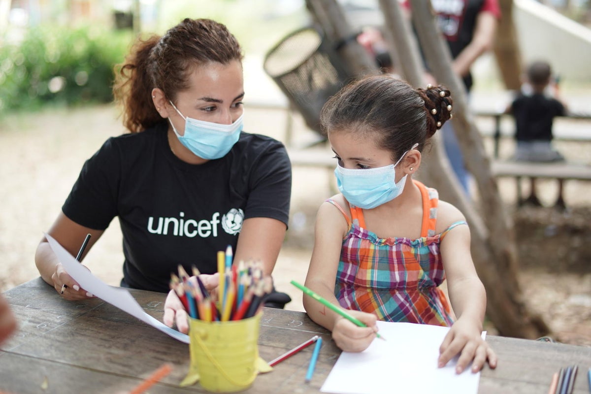 A woman with a UNICEF T-shirt is seating on a table with a young girl. The woman is showing something for the girl to draw on a piece of paper.