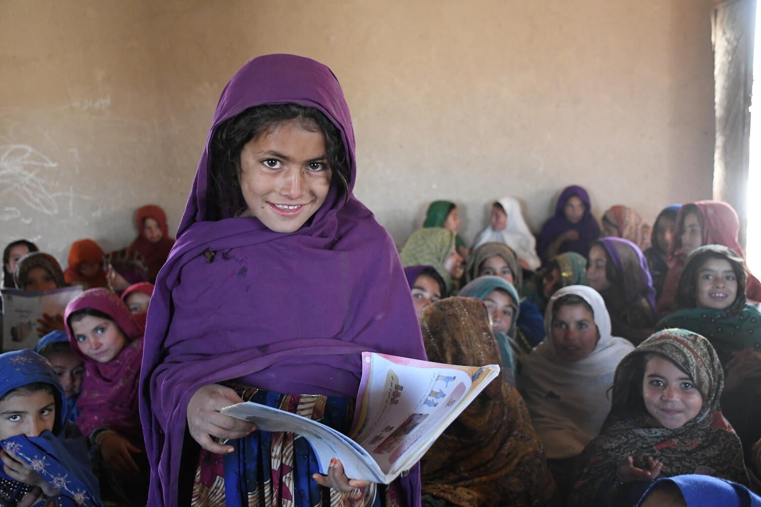 A young girls is holding a book and looking at the camera. In the background, her classmates are seating on the classroom