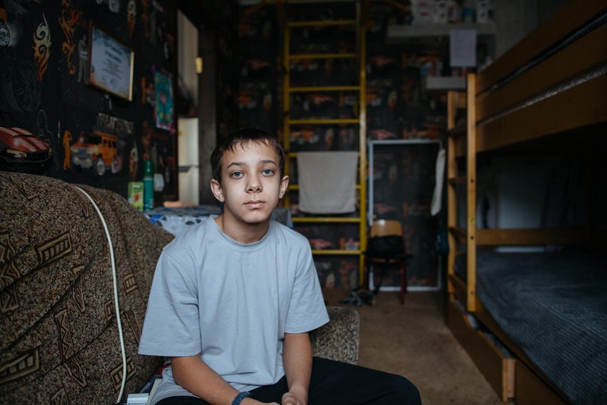Andrii sits in the communal room where his family is temporarily living in Ukraine. Bunk beds and basic furniture fill the narrow space, which the family uses for sleeping, studying and daily life.