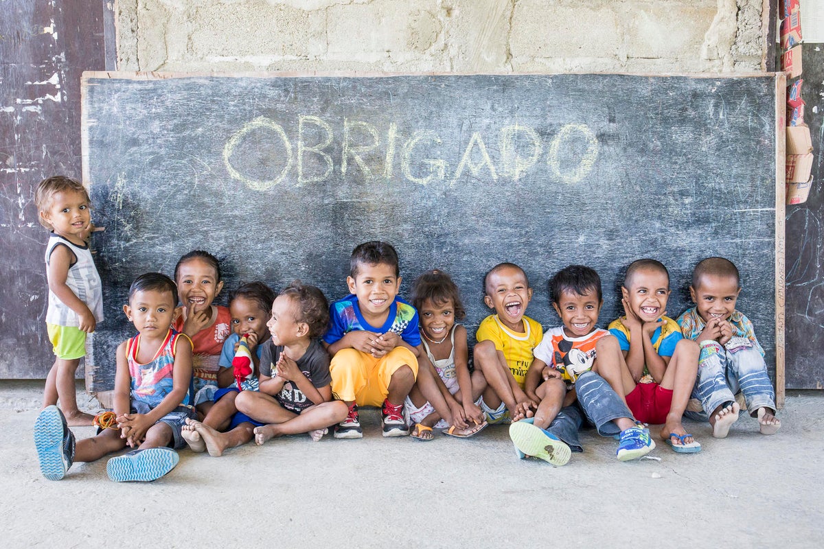 A group of 10 young children are seating on the floor smiling. Behind them, there's a blackboard that has 'Obrigado' (thank you) written on it.