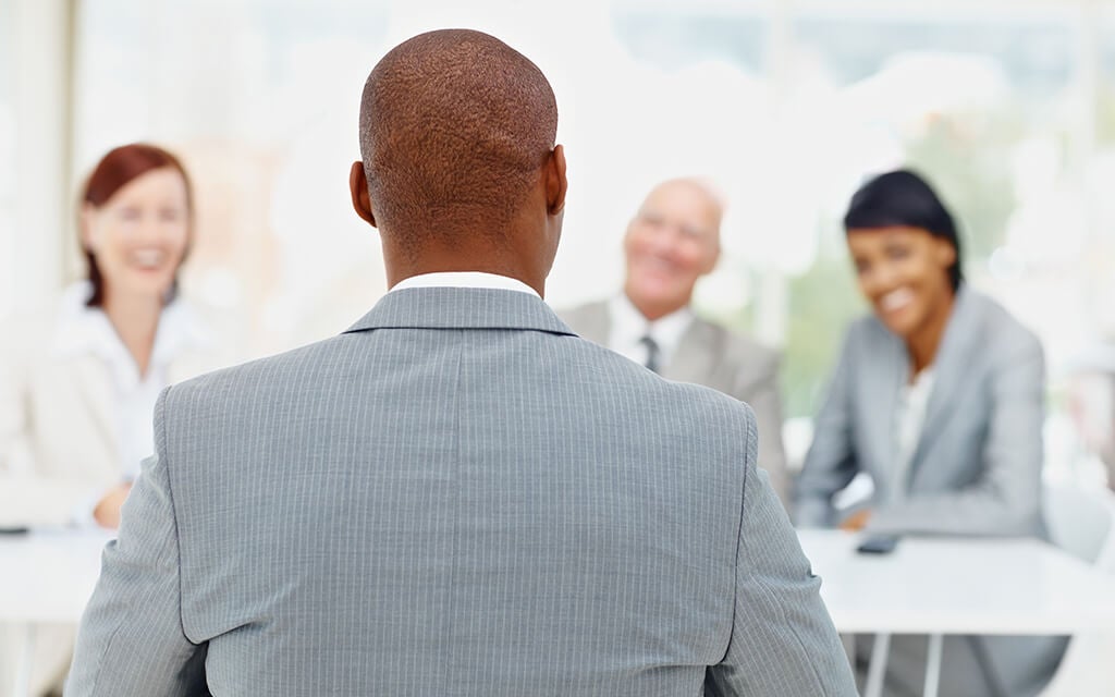 An image of a man in a suit facing away from the lens, talking to a table of people.