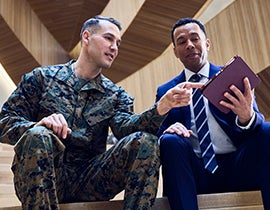 Two people seated indoors, one wearing a military uniform and the other in business attire, reviewing documents together.