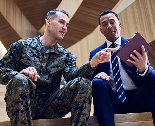 Two people seated indoors, one wearing a military uniform and the other in business attire, reviewing documents together.