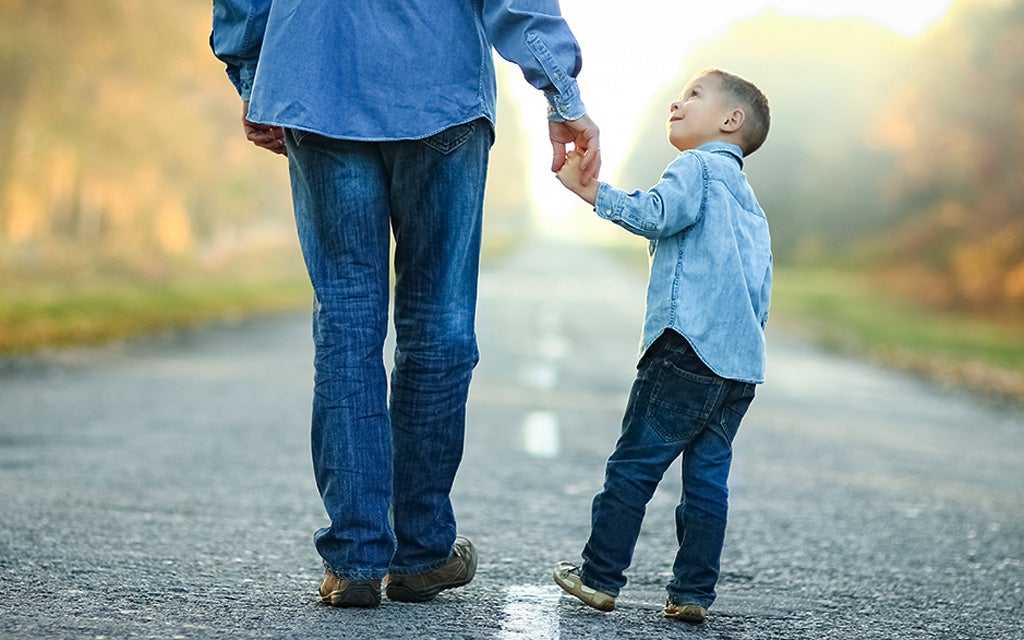 Parent walking down the street with a small child holding their hand and looking up at them.