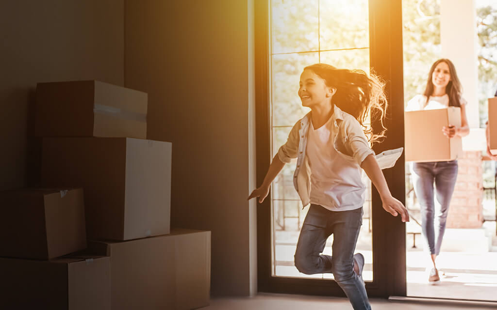 Two millennial first-time home buyers carrying moving boxes and watching as their young daughter runs into their new home.