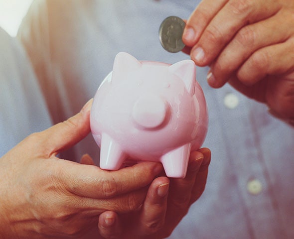 Close-up of two hands holding a pink piggy bank while another hand places a coin into it, symbolizing saving money.