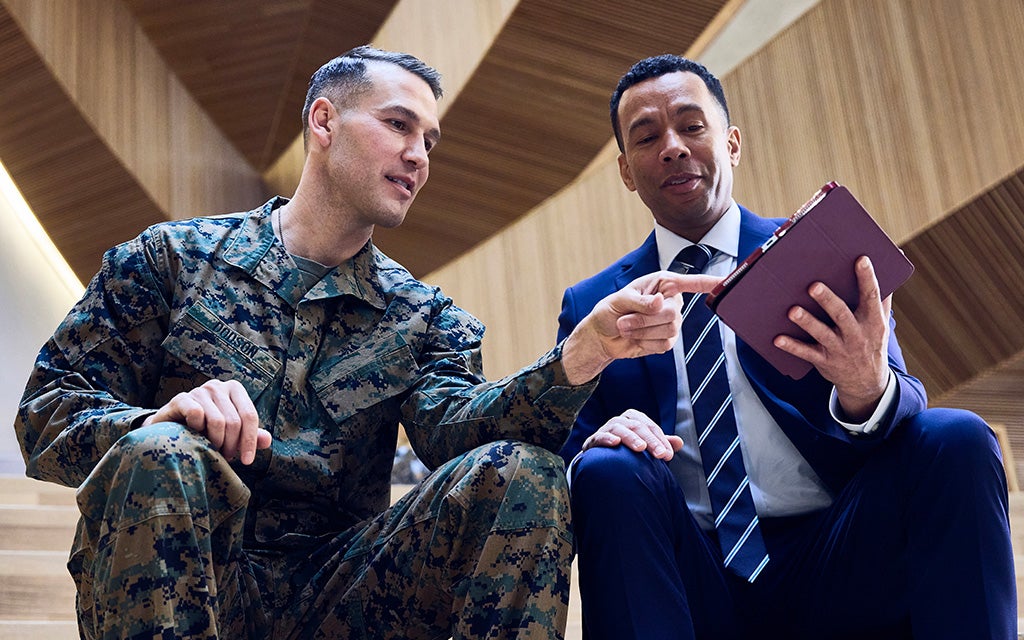 Two people seated indoors, one wearing a military uniform and the other in business attire, reviewing documents together.