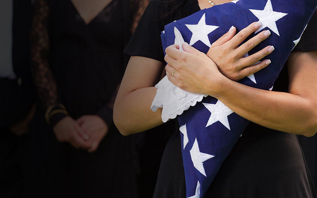 Military widow in focus holding her chest with an American flag folded inside her arms.