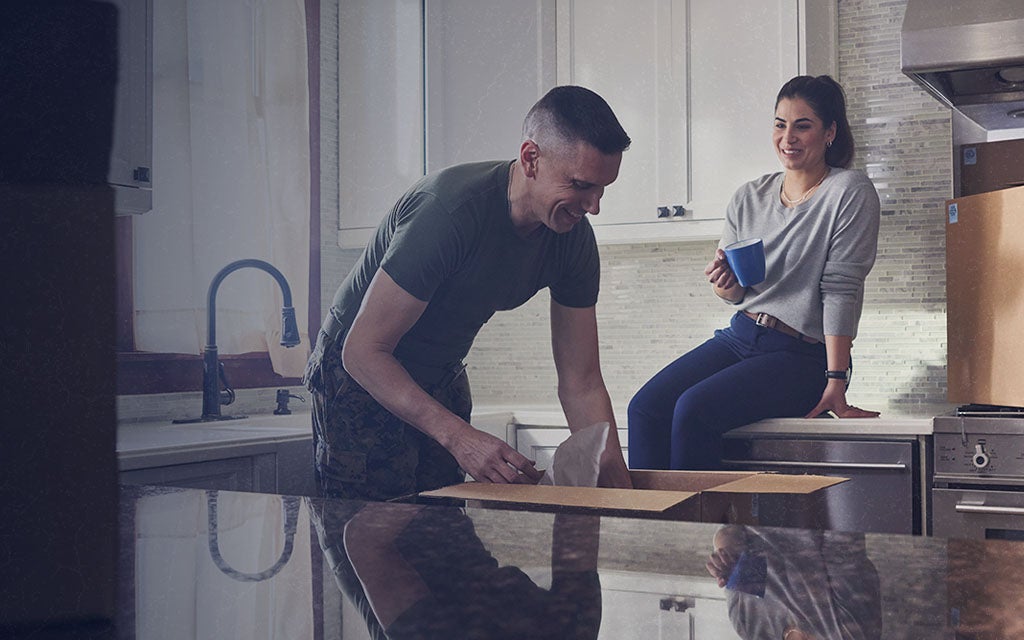 A man and a woman unpack items in a modern kitchen, with the man working at the counter and the woman sitting on the countertop holding a mug.