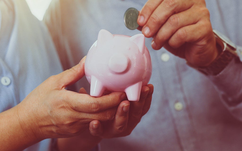 Close-up of two hands holding a pink piggy bank while another hand places a coin into it, symbolizing saving money.