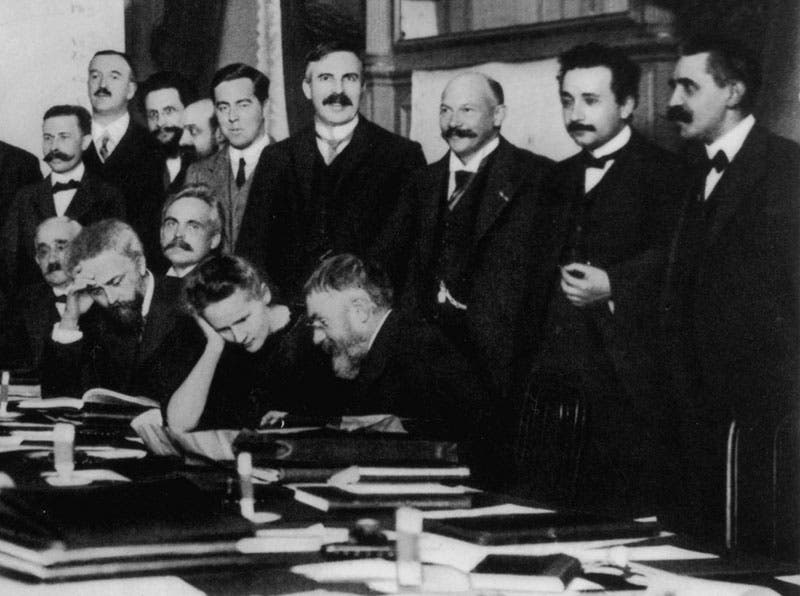 Detail of second image; Marie Curie is the only woman present, discussing a problem with Henri Poincaré; Paul Langevin is a far right, next to Albert Einstein; Jean Perrin is seated just to the left of Madame Curie, reading a book (Wikimedia commons)
