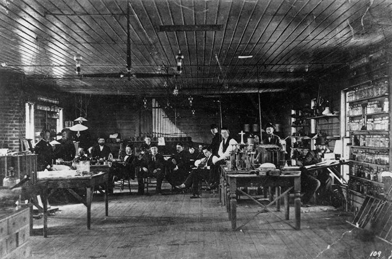 Interior of one of the original Menlo Park Laboratory buildings, Thomas Edison seated at left-center, in skull cap, with workers and staff gathered around, photograph, late 1870s, Emilio Segrè Visual Archives, American Institute of Physics (repository.aip.org)