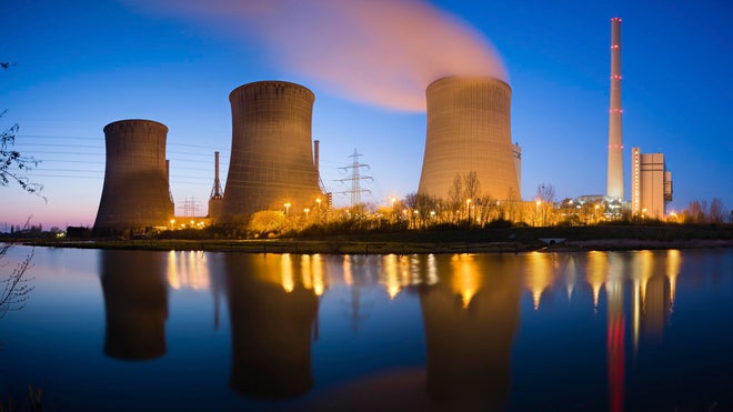 A power plant with four cooling towers emitting steam and a tall chimney, all reflected on a calm body of water during twilight