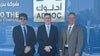 Three men, one in sunglasses, standing against ADNOC wall with English and Arabic writing