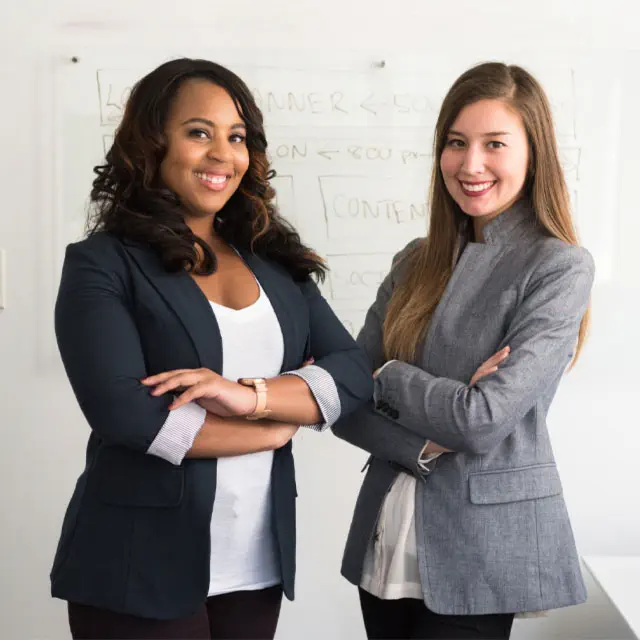 Two business women proudly standing shoulder-to-shoulder in front of a whiteboard in a conference room.