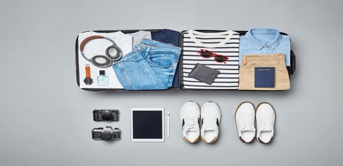 Maleta abierta con ropa doblada y artículos de viaje como auriculares, cámaras, perfumes, zapatos, tablet y pasaporte, organizada sobre un fondo gris.