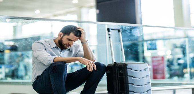 Hombre con gesto preocupado sentado junto a su maleta en sala de espera de aeropuerto.