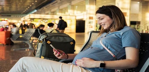 Mujer embarazada sentada en el aeropuerto revisando su celular mientras espera su vuelo.