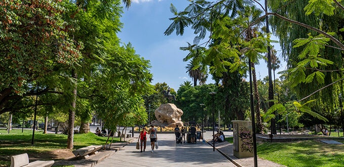 Sendero arbolado en Plaza San Martín, Mendoza, con personas paseando y una gran escultura rocosa al centro.