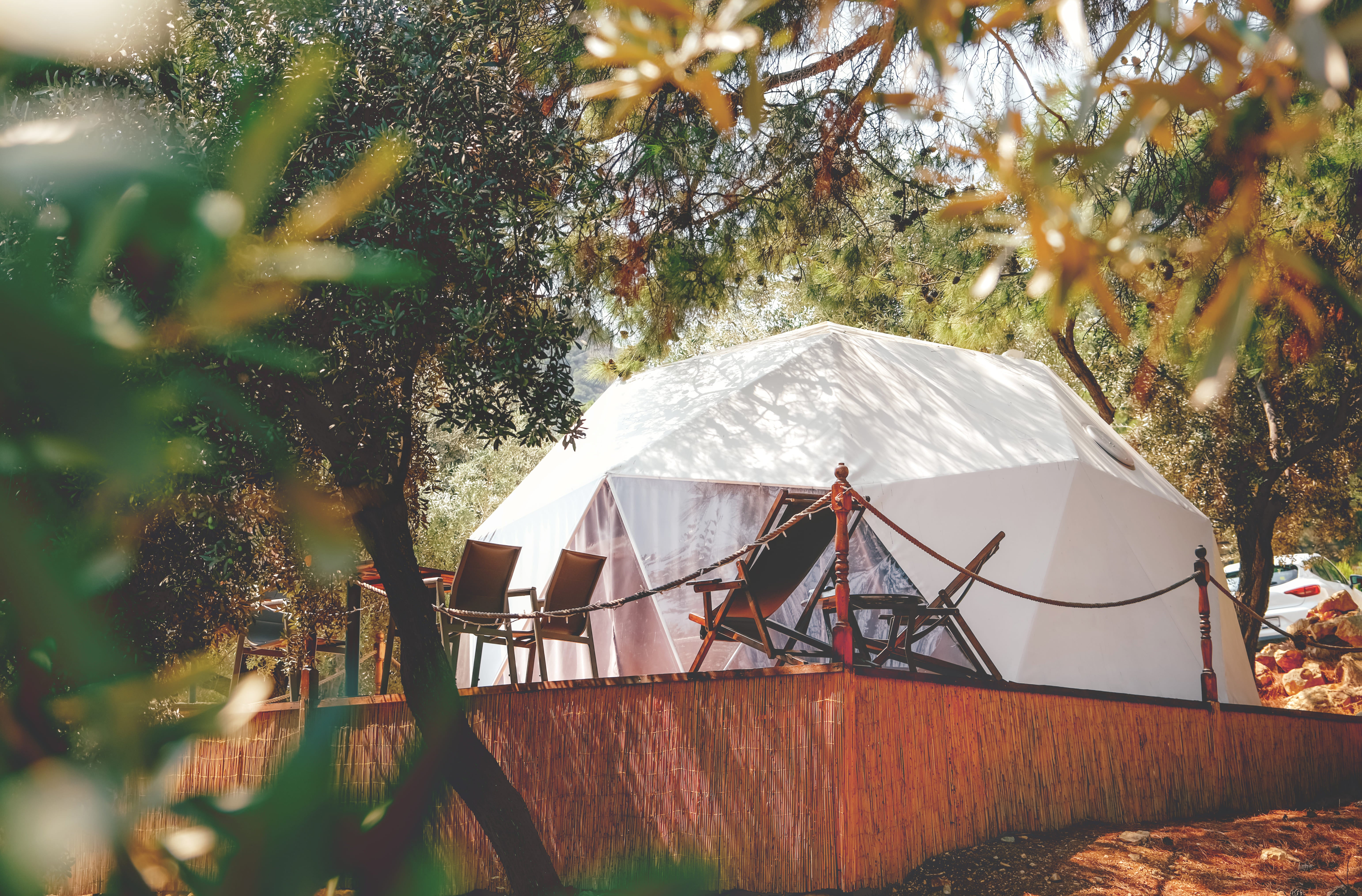 Domo de glamping blanco en plataforma de madera, rodeado de árboles en entorno boscoso y soleado.