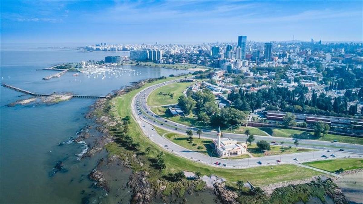 Vista aérea panorámica de la costa de Montevideo con la ciudad al fondo, destacando su rambla y zonas verdes.
