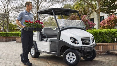 A man places a large case of flowers onto the back of a Club Car electric utility vehicle on a college campus.