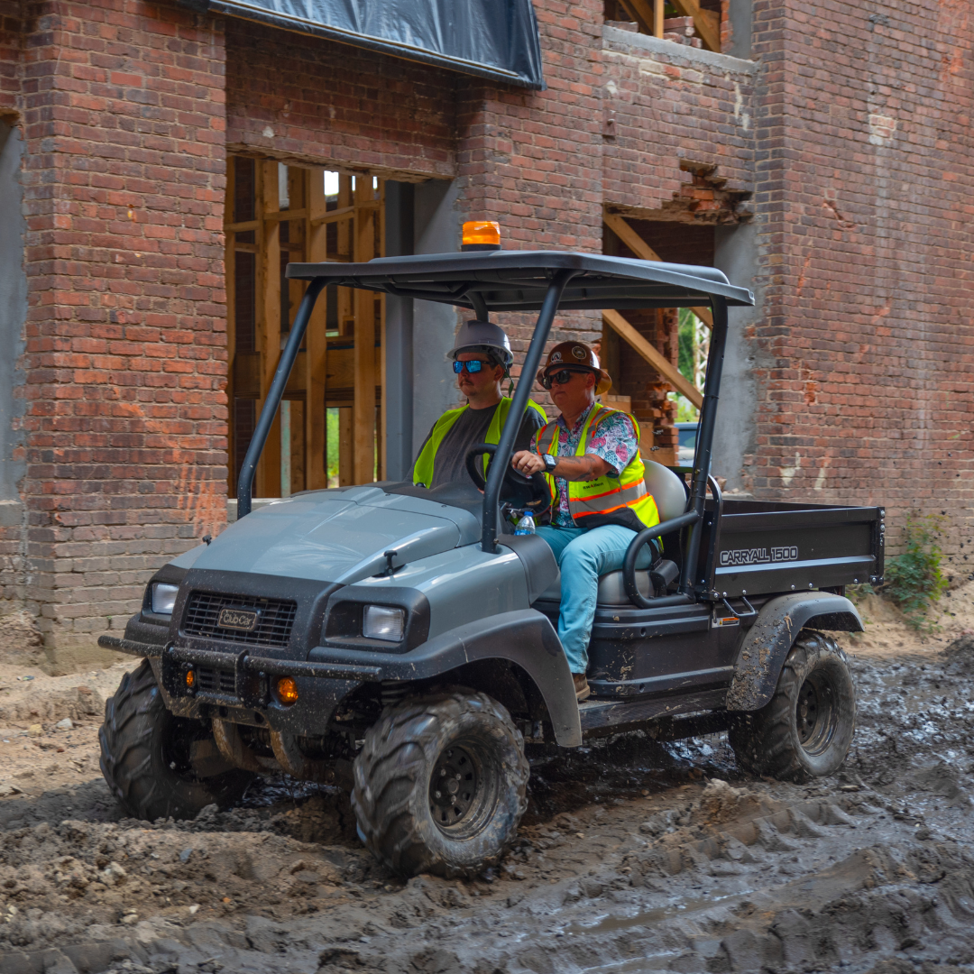 Two men in construction gear drive a Club Car Carryall utility vehicle through a construction zone.