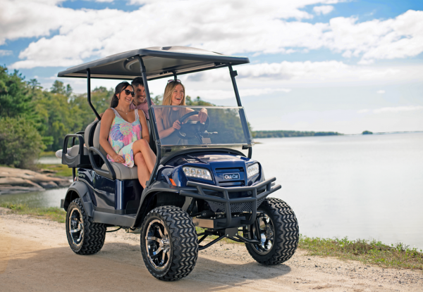 Three people drive a Club Car golf cart along a lakefront.