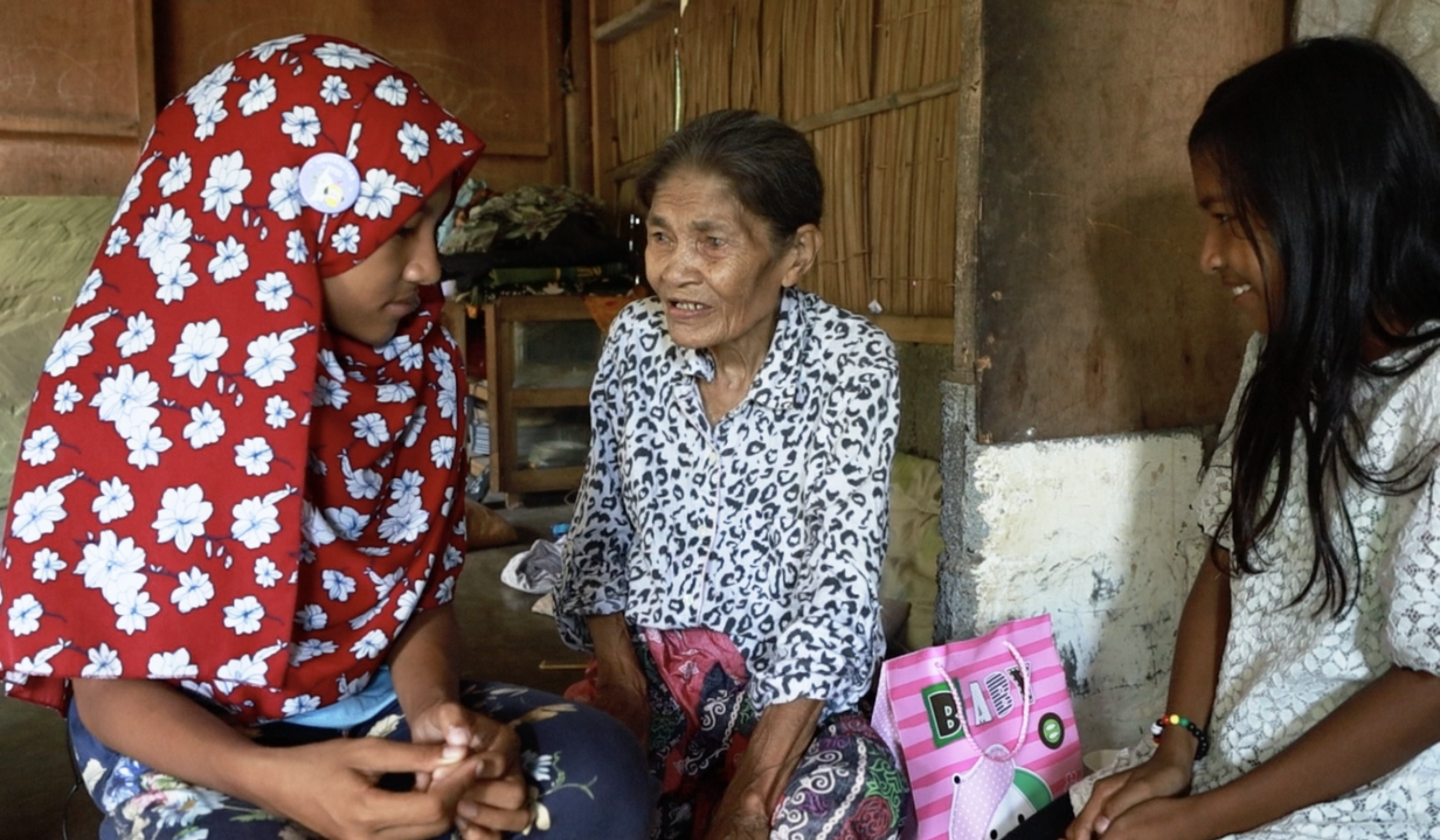 In the Philippines, two young sisters with their grandmother. One of the girls wears a red floral head covering. 