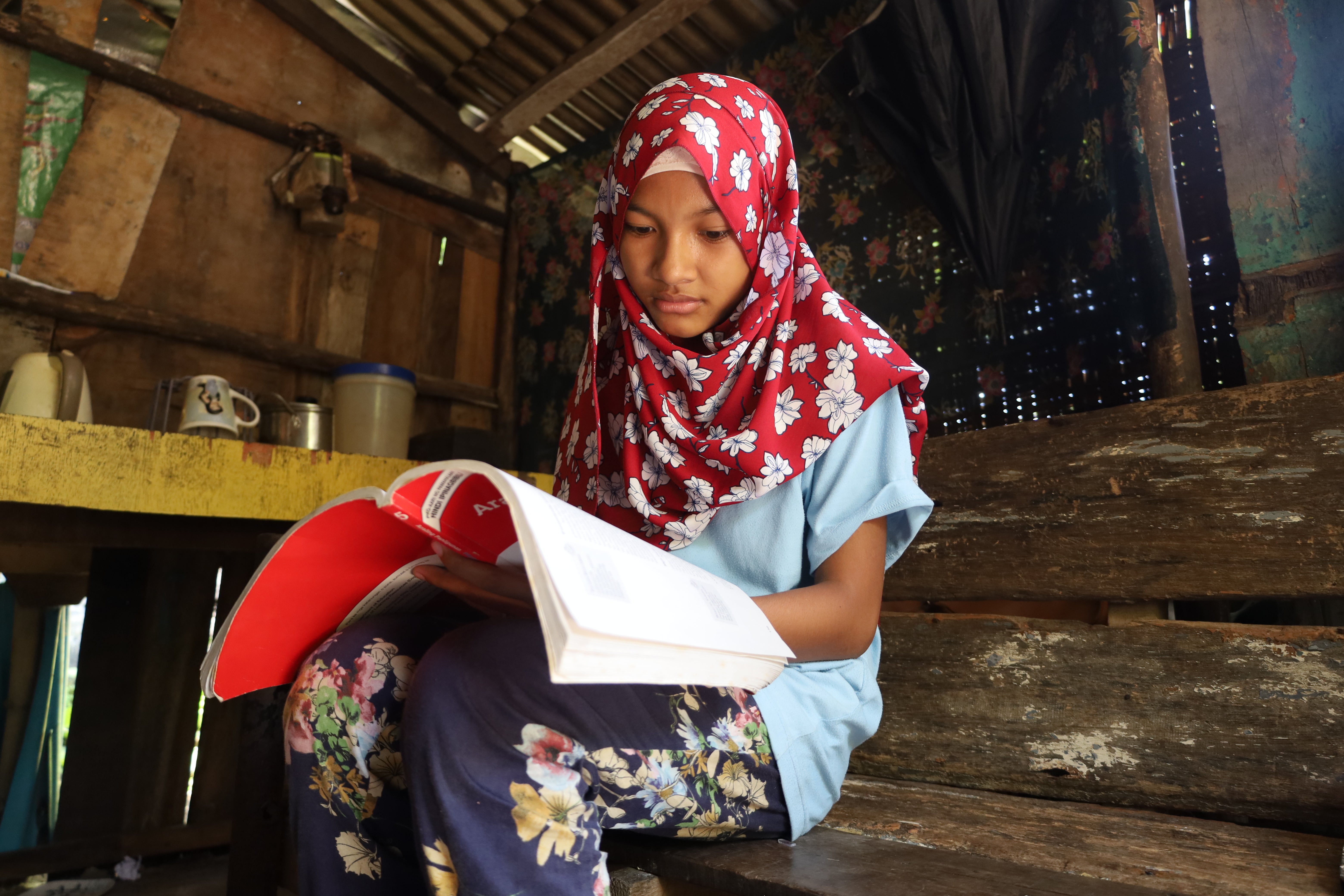 A young girl wearing a red floral head covering sits inside on a wood bench and reads a textbook. 