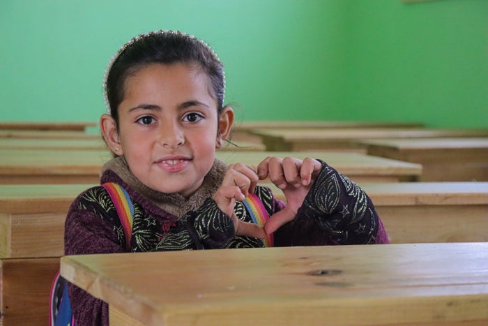 In Syria, a young girl smiles and makes a heart symbol with her hands as she sits at a school desk. 