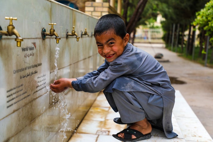 In Afghanistan a young boy crouches by a water tap wall and holds his hands under the running water. He smiles for the camera.