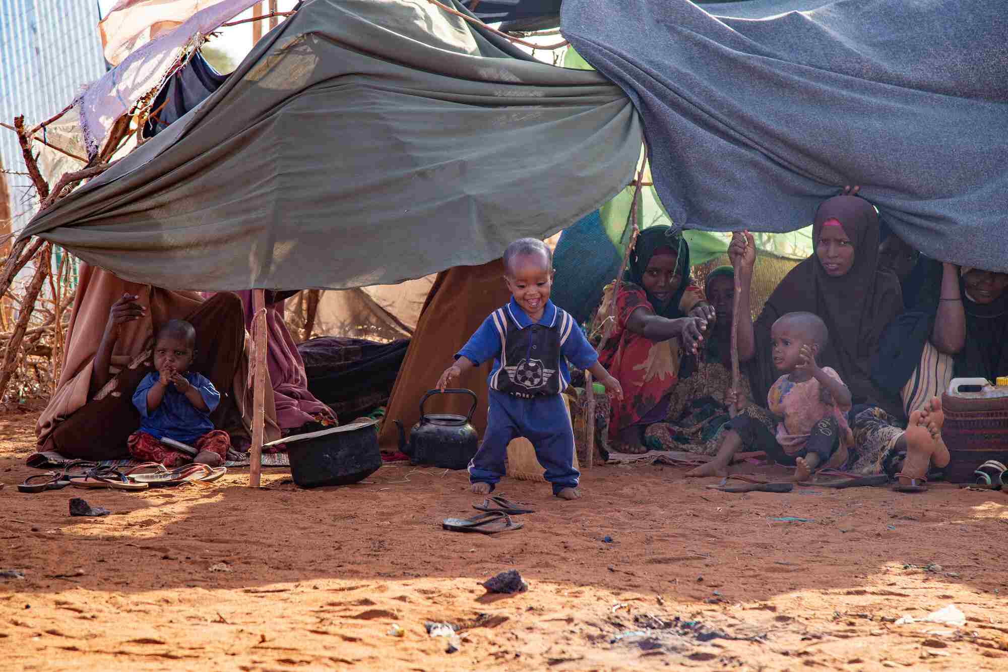 A toddler boy smiles as he walks out of a makeshift shelter. Women and children are seen in the shelter behind him.