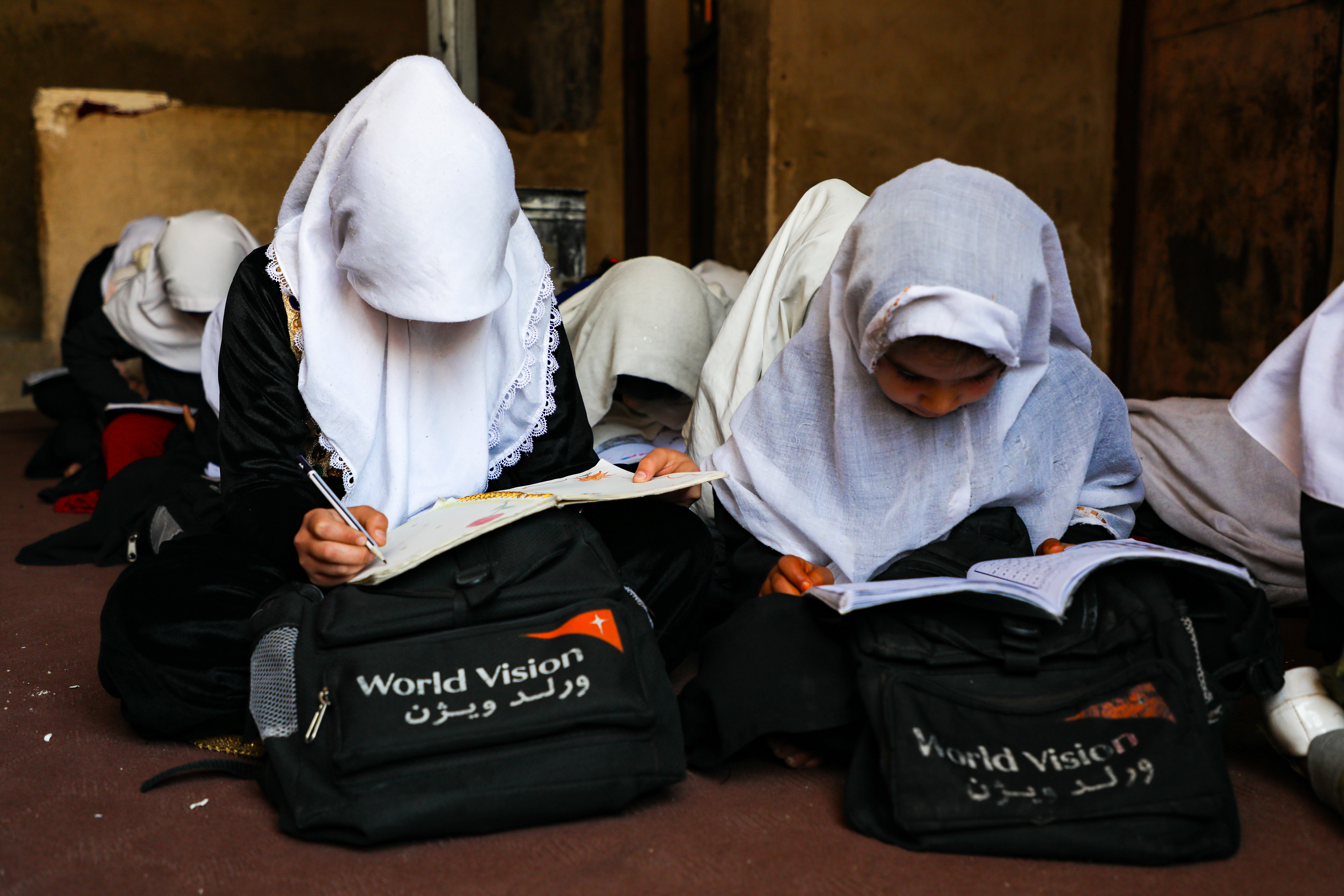 In Afghanistan, two girls in head coverings sit on the floor in a classroom working in their notebooks. They each have a World Vision Afghanistan backpack. 